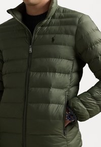 Mężczyzna w oliwkowo-zielonej pikowanej kurtce z zamkniętą kieszenią i ciemnym zegarkiem widocznym na nadgarstku.