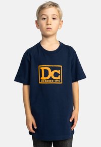 Marinblå bomullströja med en stor orange DC Shoes-logotyp och text. Korta ärmar och en avslappnad passform, med rund halsringning.