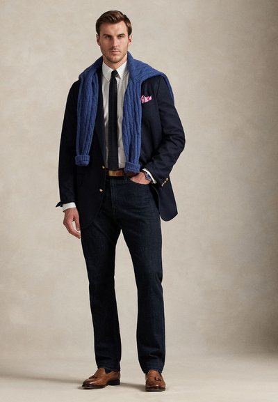 Man wearing navy blazer, white shirt, black tie, dark jeans, brown loafers, and blue sweater draped over shoulders, standing against plain background.