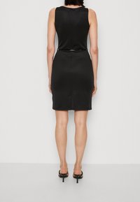 Robe noire sans manches avec une silhouette ajustée, fermeture éclair à l'arrière et une ceinture noire brillante soulignant la taille.