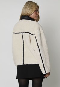 Cremefarvet fluffy jakke med sort læderkrave, med sorte piping-kanter og en boxy form. Rygview af modellen.