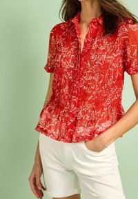 Blouse courte à manches courtes rouge avec un motif floral, texture plissée et un ourlet en peplum. Associée à un short blanc, sur un fond vert clair.
