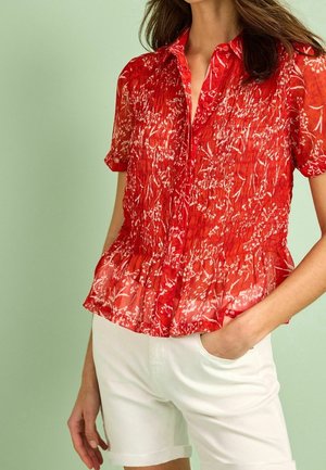 Blouse courte à manches courtes rouge avec un motif floral, texture plissée et un ourlet en peplum. Associée à un short blanc, sur un fond vert clair.