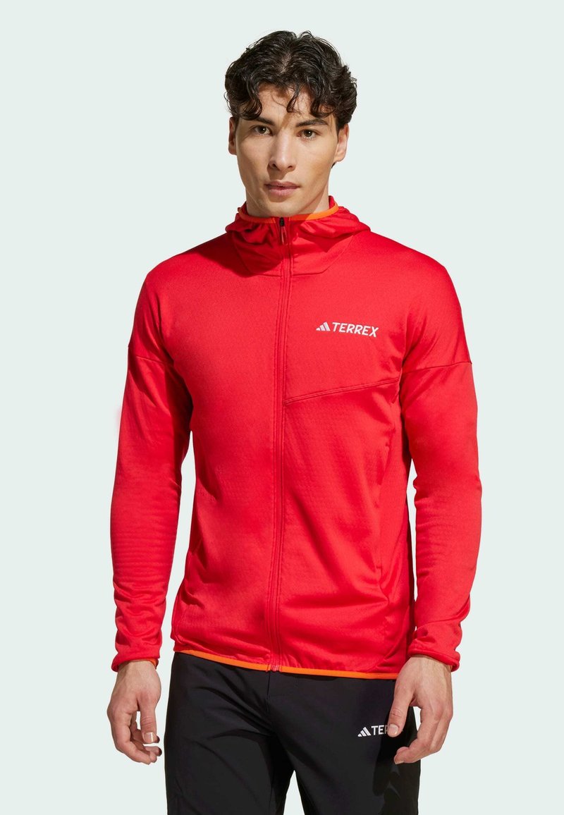 Red zip-up athletic jacket with a textured finish, featuring a hood, long sleeves, and a logo on the chest. Orange accents at the hem.