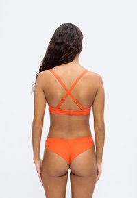 Γυναίκα με μακριά σκούρα μαλλιά που φοράει πορτοκαλί μπικίνι με χιαστί τιράντες στην πλάτη και λογότυπο Puma στο κάτω μέρος, στέκεται μπροστά σε απλό φόντο.