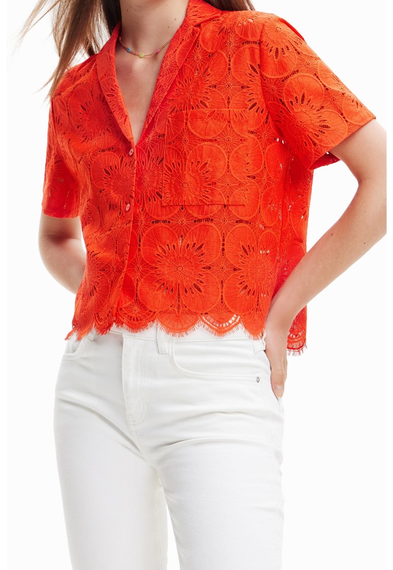 Camicia rossa in pizzo a maniche corte con motivi floreali, chiusura con bottoni davanti, orlo a scallop e taschino sul petto; indossata sopra pantaloni bianchi.