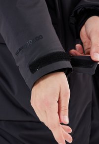 Svart vattentät jackärm med justerbart kardborrefäste, med "W-PRO 10000" tryckt i svart på tyget. Slät textur.