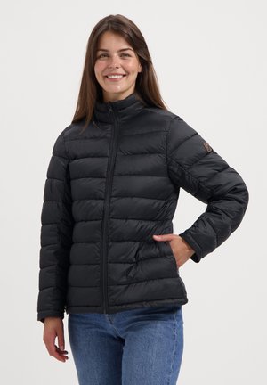 Femme souriante portant une veste matelassée noire et un jean bleu se tient avec une main dans la poche contre un fond clair et uni.