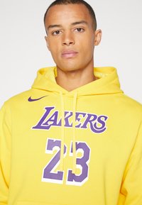 Moletom amarelo de algodão com um bolso canguru na frente. Apresenta o texto roxo "LAKERS" e o número "23" em branco com um padrão de pontos.