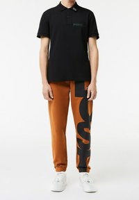 Polo noir avec détails en texte vert, associé à un pantalon de survêtement orange avec de grandes lettres noires. Des baskets blanches complètent le look.