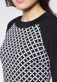 Sweter w czarno-białe wzory z geometrycznym designem, z czarnym żebrowanym dekoltem i kontrastującą fakturą w rękawach.
