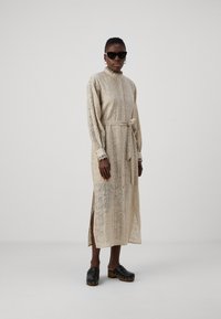 Beige, long-sleeve dress with a patterned texture, button-front, tied waist, and side slits. Paired with black clogs featuring gold accents.