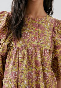 Robe fleurie aux tons jaunes et roses, avec des manches bouffantes, un col rond et des boutons en décoration. Le tissu présente un motif texturé et travaillé.