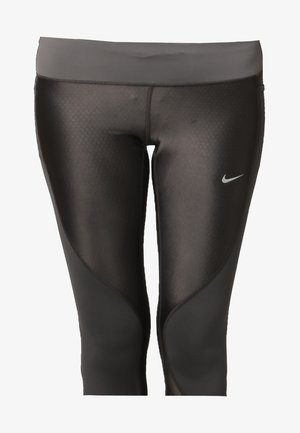 Kobieta w czarnym sportowym topie bez rękawów, czarnych krótkich legginsach z logo Nike i czarnych butach do biegania, stojąca na jednolitym tle.