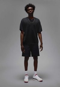 Camicetta nera a maniche corte con texture, pantaloni corti neri, sneakers bianche con dettagli rossi, indossate con calzini bianchi fino alla caviglia. Collana semplice a catena.
