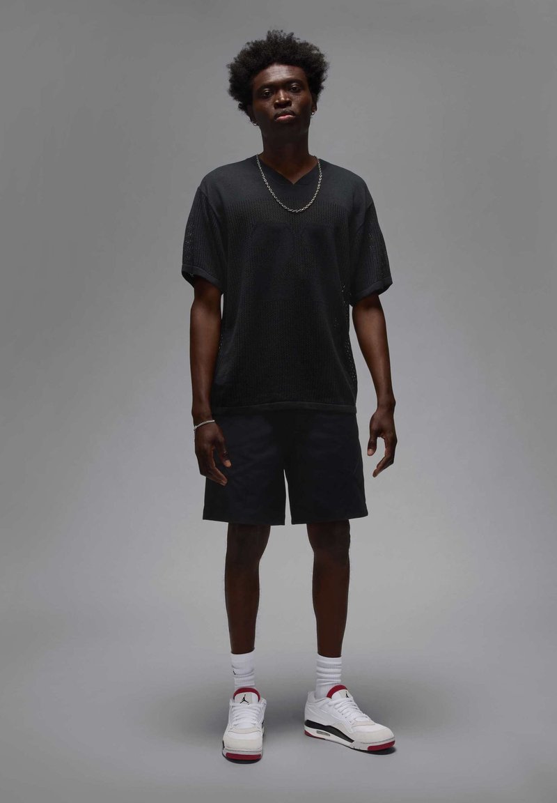 Camicetta nera a maniche corte con texture, pantaloni corti neri, sneakers bianche con dettagli rossi, indossate con calzini bianchi fino alla caviglia. Collana semplice a catena.