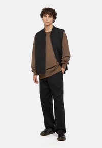 Chaleco negro sin mangas sobre una camisa de manga larga marrón, combinado con pantalones negros holgados y zapatos negros. Tela suave con detalles mínimos.