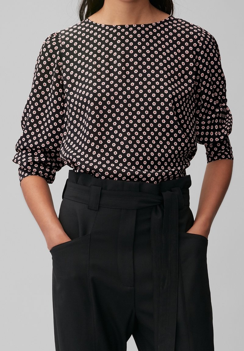 Femme portant un chemisier noir à motifs de fleurs roses et un pantalon noir taille haute avec une ceinture en tissu, les mains dans les poches.