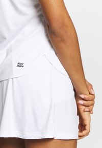 Vit sporttopp i tanktop-stil med mesh-textur, med rundad nederkant och varumärkeslogotypen "BIDI BADU" på sidan. Matchas med en vit kjol.