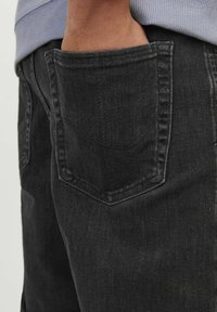 Jean noir en denim avec une finition texturée, comprenant une poche arrière. Les coutures sont discrètes et le tissu présente une légère usure.