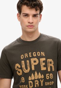 Camiseta de algodón verde olivo oscuro con un estampado gráfico texturizado en amarillo que dice "OREGON SUPER DRY" y motivos de árboles. Mangas cortas y cuello redondo.
