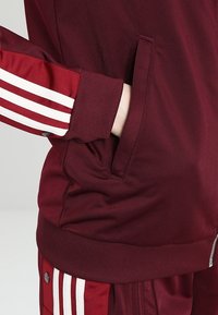 Bordeauxrode sportjas met witte strepen op de mouwen; voorzien van zijzakken en geribbelde manchetten; gladde textuur en een aansluitend ontwerp.