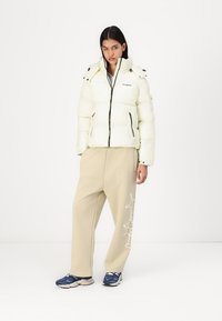 Pufferjacke in lichtgeel, met een ritssluiting aan de voorkant, dubbele schouderknopen en zwarte accenten; gecombineerd met beige broeken en marineblauwe sneakers.