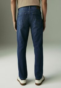 Blugi din denim albastru au un croi drept, două buzunare la spate cu detalii de cusătură și un patch din piele cu logo la talie.
