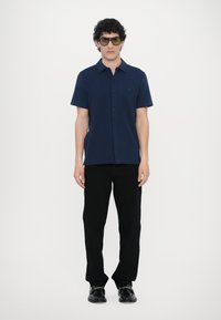 Navy short-sleeve button-up shirt made of smooth fabric, paired with black trousers and black shoes with decorative hardware.
