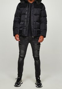 Mand iført en sort polstret jakke med pelskrave, slidte sorte skinny jeans og sort-hvide sneakers, stående mod en ensfarvet baggrund.