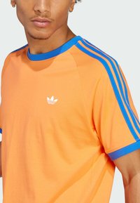 Camiseta de algodón naranja con acentos azules en el cuello y las mangas, con tres rayas azules en cada hombro y un logo blanco en el pecho.