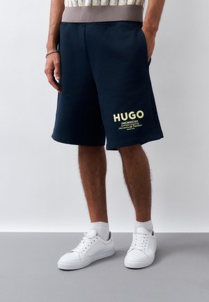 Άνδρας που φοράει σκούρο μπλε σορτς Hugo με κίτρινο λογότυπο στο αριστερό πόδι, λευκά αθλητικά παπούτσια και λευκές κάλτσες, στέκεται μπροστά σε απλό φόντο.