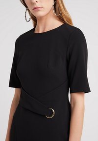 Svart klänning med korta ärmar, med en insvept design och en guldring som accent vid midjan. Slätt tyg med en strukturerad form.