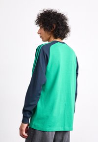 Green and navy long-sleeve shirt with raglan sleeves. Features three green stripes on the shoulders and a rounded neckline. Smooth texture.