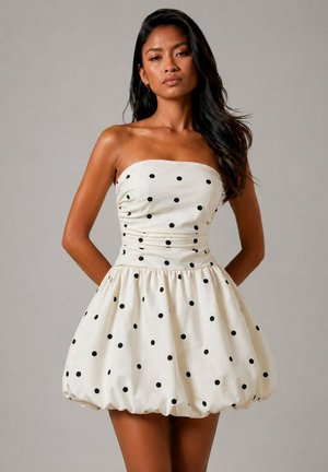 Femme portant une robe blanche bustier bouffante avec des pois noirs, debout les mains dans le dos sur un fond gris uni.