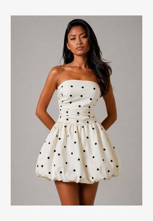Femme portant une robe blanche bustier bouffante avec des pois noirs, debout les mains dans le dos sur un fond gris uni.