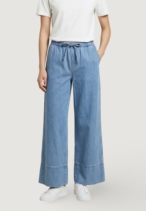 Persona vistiendo pantalones de mezclilla azul claro de pierna ancha con cintura elástica y cordón, zapatillas blancas y una camiseta blanca lisa de manga corta.