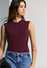 Top sin mangas color burdeos con cuello alto, combinado con jeans de mezclilla azul claro. El top tiene una textura suave y un diseño ajustado.