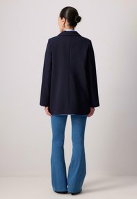 Granatowa marynarka o dopasowanym kroju, szerokich klapach i gładkiej teksturze, w zestawieniu z dzwonami z niebieskiego denimu. Czysty design, bez widocznych wzorów.