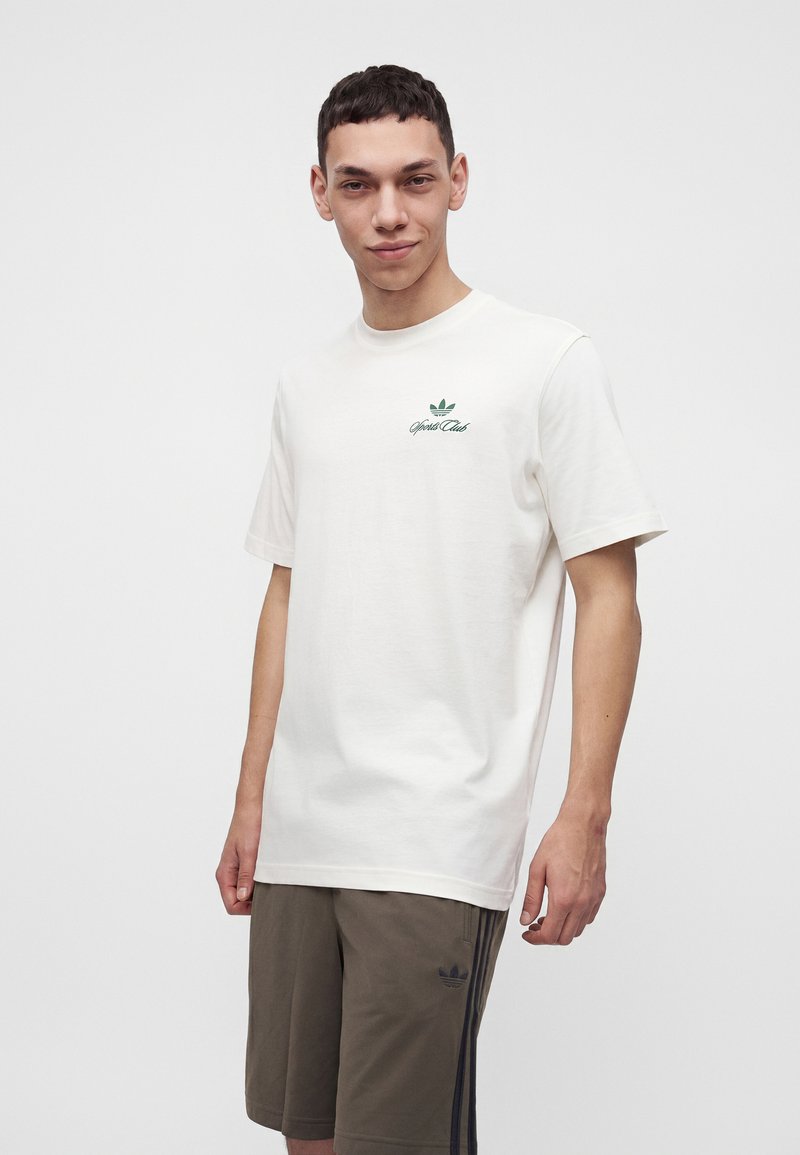 Junger Mann trägt ein weißes kurzärmliges T-Shirt mit grünem Logo und olivgrüne Shorts mit schwarzen Streifen vor einem einfachen weißen Hintergrund.