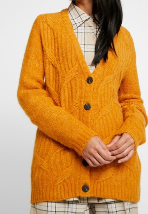 Žena v hořčicově žlutém kardiganu s copánkovým vzorem a černými knoflíky, přes bílou kostkovanou košili a v odpovídající sukni, s rukama sepjatýma.