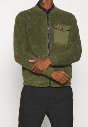 Mand iført olivengrøn tekstureret jakke med lynlås og brystlomme samt sorte bukser, armene krydset ved taljen.