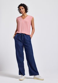 Różowa, bezrękawnikowa bluzka z dekoltem w kształcie litery V, zestawiona z luźnymi, ciemnoniebieskimi dżinsami i białymi trampkami. Materiał wydaje się gładki o lekkiej teksturze.