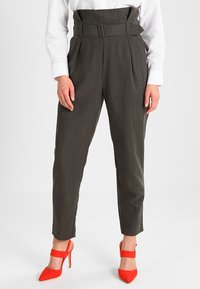 Kobieta nosząca ciemnoszare spodnie z wysokim stanem, z materiałowym paskiem i jasnoczerwonymi czółenkami na wysokim obcasie z zapięciem wokół kostki.