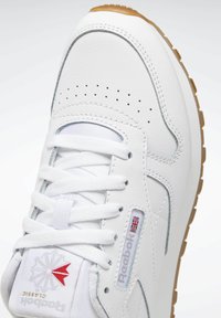 Zapatilla de cuero blanca con punta redondeada, detalle perforado, cordones blancos y suela de goma. Presenta una etiqueta de logotipo con un acento rojo.