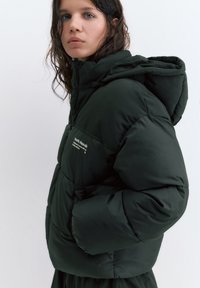 Mladá osoba s mokrými vlasmi, oblečená v čiernej páperovej bunde s kapucňou, stojaca bokom na plainom bielom pozadí.