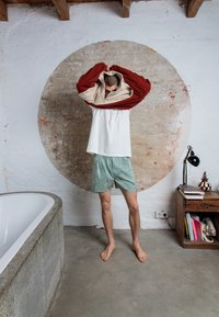 Une personne portant un pull en polaire beige et rouge avec fermeture éclair, une chemise blanche et un short vert à motifs se tient dans une pièce minimaliste avec une baignoire et une lampe.