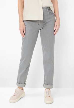 Person iført lysegrå bukser med opbuk, beige sneakers og en beige kortærmet top, stående mod en hvid baggrund.