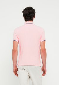 Ροζ πόλο πουκάμισο με εφαρμοστή σχεδίαση, κοντά μανίκια και αντίθετη σκούρα επένδυση για το γιακά. Το ύφασμα φαίνεται λείο, ελαφρύ και διαπνέον.