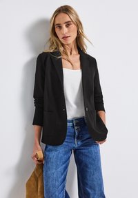 Černý blazer s jedním knoflíkem, dvěma předními kapsami a dlouhými rukávy, kombinovaný s bílým topem a modrými džínami.